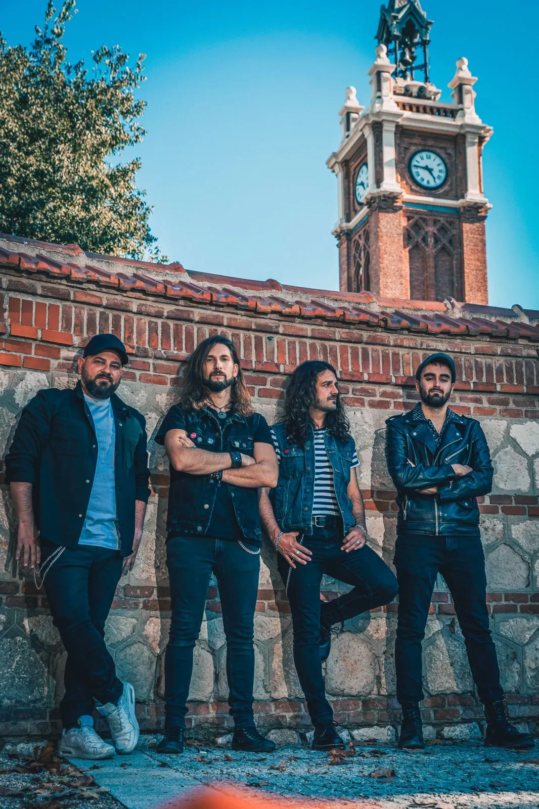 Los cuatro músicos de A TiRO junto a un muro de ladrillo y una torre con reloj
